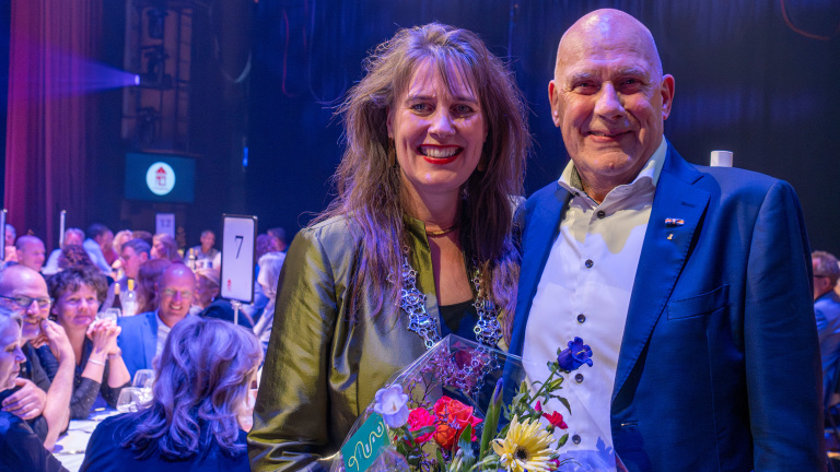 Twee mensen poseren samen op een evenement met een publiek op de achtergrond. De vrouw houdt een boeket bloemen vast, terwijl de man een donkerblauw pak draagt. Ze glimlachen beiden naar de camera.