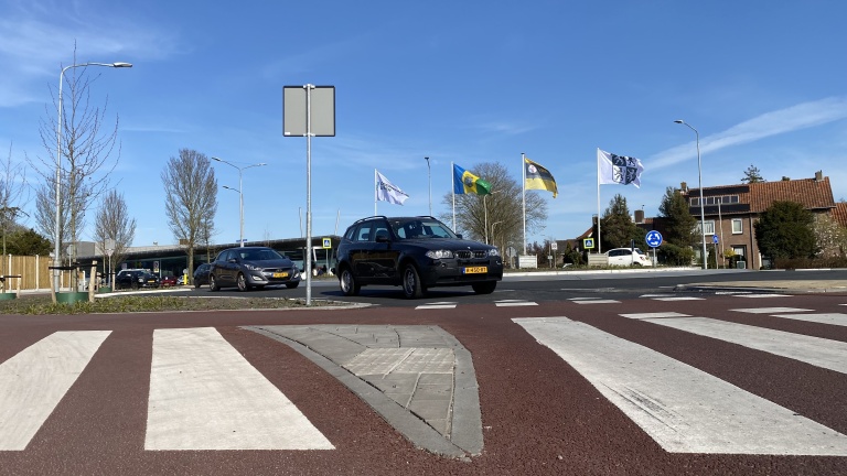 Auto's rijden op een rotonde met zebrapaden en verschillende vlaggen aan masten in de achtergrond, onder een heldere blauwe lucht.