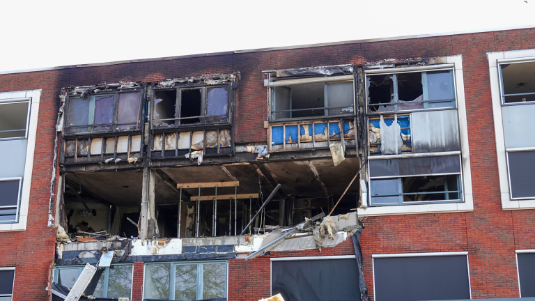 Een zwaar beschadigde gevel van een appartementencomplex, met uitgebrande en ingestorte verdiepingen, zichtbaar zwartgeblakerde muren en kapotte ramen.