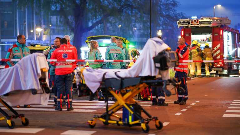 Hulpverleners in uniform staan 's avonds bij een ambulance en brandweerauto op een weg, met brancards op de voorgrond.