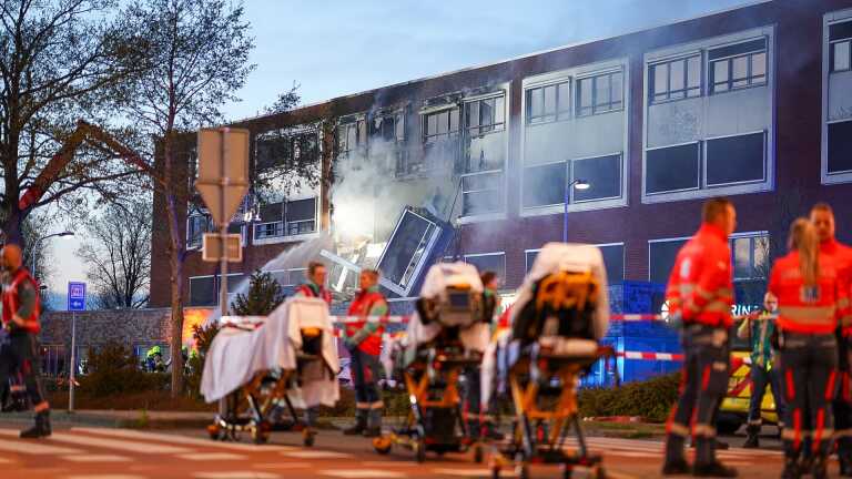 Ambulancepersoneel en brancards voor een gebouw dat schade en rookontwikkeling vertoont na een explosie.