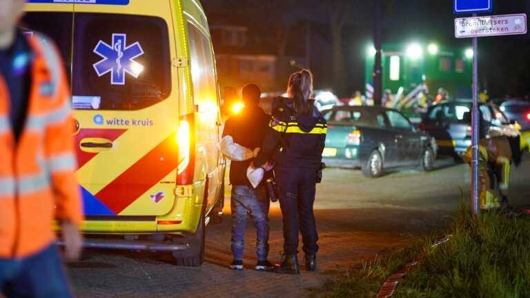 Een agent ondersteunt een jongen richting een ambulance in de nacht, omringd door geparkeerde auto's en nooddienstpersoneel.
