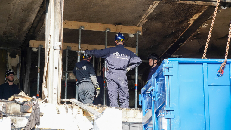 Forensisch politiepersoneel onderzoekt een beschadigd gebouw, met metalen stutten en een blauwe container op de voorgrond.