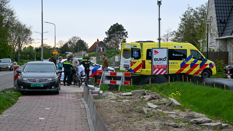Een ambulance en politieagenten op een trottoir naast een weg, met een geparkeerde Honda en een groep mensen die omstanders lijken te zijn.