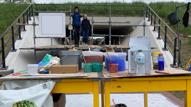 Twee personen staan achter een tafel met verschillende potten verf en materialen op een bouwplaats bij een tunnelconstructie, omgeven door hekken en groen gras.