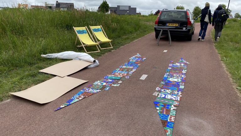 Twee gele strandstoelen en kartonnen stukken liggen op een fietspad naast een rode auto. Op het pad liggen kleurrijke, met patronen beschilderde stukken papier of stof in de vorm van pijlen. Twee personen lopen verderop naast de auto.