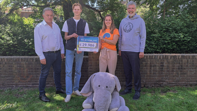 Vier mensen poseren buiten voor een bakstenen muur met een groot cheque-bord waarop €29.986 staat; een grote knuffelolifant zit op de grond.