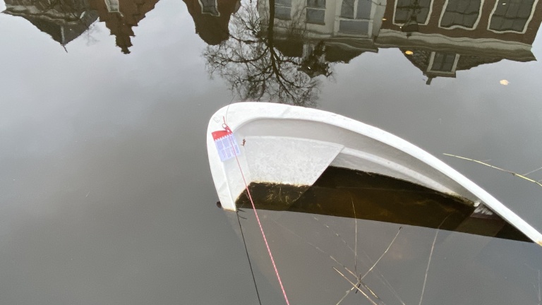 Een half gezonken wit bootje in een rustige gracht, met op de achtergrond de reflectie van huizen en bomen in het water.