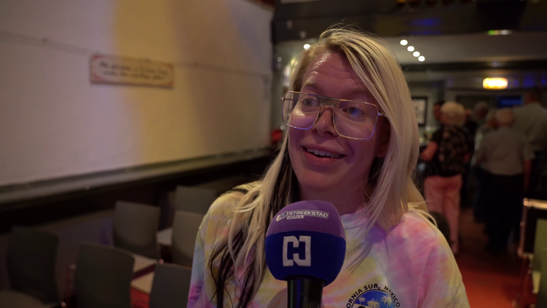 Een vrouw met lang blond haar en een bril spreekt in een microfoon met het logo van "Streekstad" erop, in een zaal met stoelen en mensen op de achtergrond.
