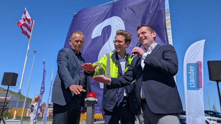 Drie mannen in formele kleding poseren lachend bij een grote rode knop op een buitenlocatie, met vlaggen van Liander op de achtergrond onder een heldere blauwe hemel.
