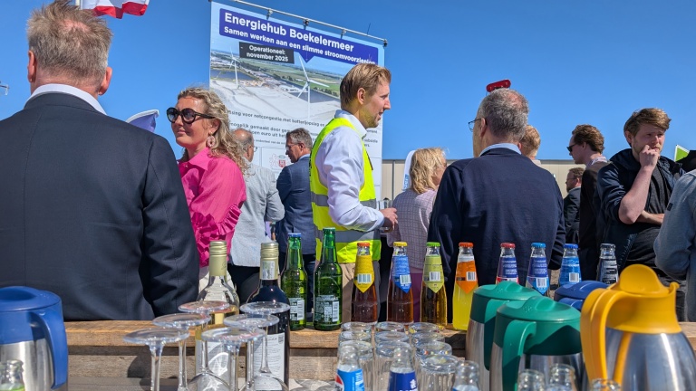 Mensen in gesprek tijdens een bijeenkomst bij Energiehub Boekelermeer, met op de voorgrond flesjes drank en glazen, en een informatiebord over een energieproject op de achtergrond.