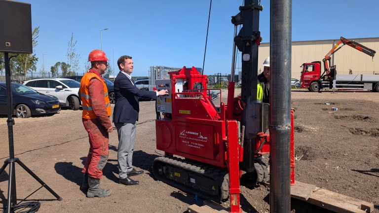 Twee mannen bedienen een rode funderingsmachine op een bouwterrein, waarbij één man in een oranje werkpak en veiligheidshelm staat en de ander in een pak, terwijl op de achtergrond auto's en een vrachtwagen zichtbaar zijn.