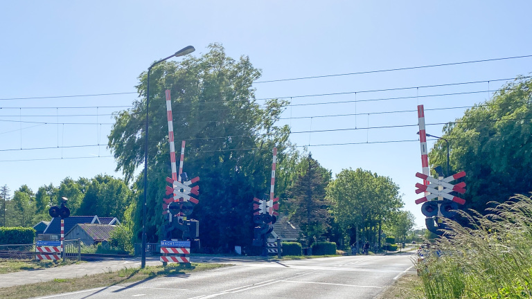Een spoorwegovergang met opstaande rode en witte slagbomen en waarschuwingslichten, omringd door groene bomen en struiken.