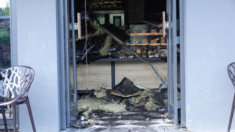 Interieur van een winkel met brandschade, inclusief gebroken plafondplaten, puin op de vloer en gedeeltelijk verbrande producten in de vitrinekast.