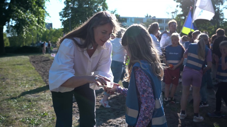 Een vrouw met een witte blouse geeft een meisje iets in haar hand terwijl ze buiten staan, omringd door andere kinderen en volwassenen.
