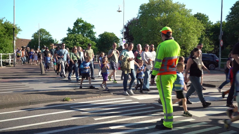 Een groep mensen, waaronder kinderen, steekt een straat over op een zebrapad, begeleid door een persoon in een felgroene veiligheidsoutfit.