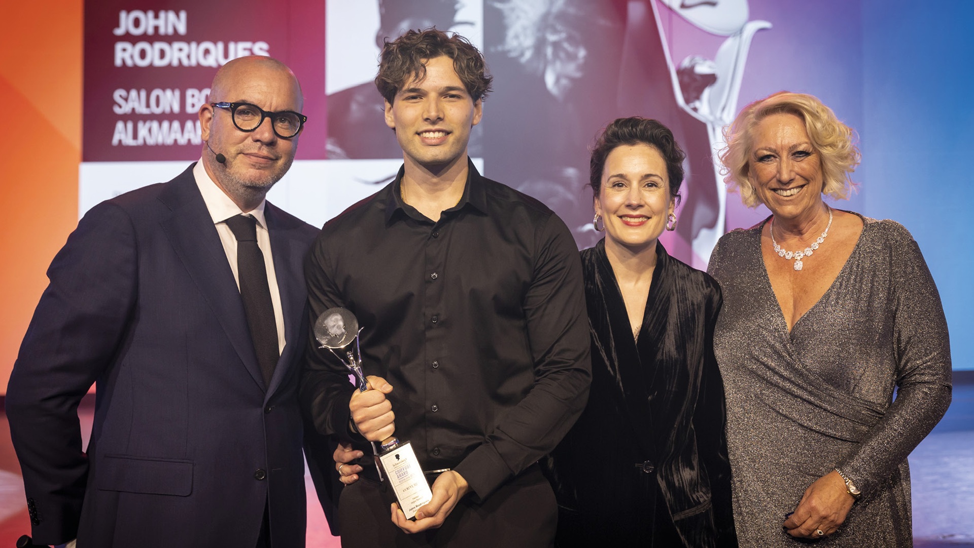 Vier mensen poseren samen op een podium; een jonge man in zwart kleding houdt een trofee vast, geflankeerd door drie andere personen, waaronder een man in pak en twee vrouwen gekleed in elegante avondkleding, met een kleurrijke achtergrond die een logo en tekst bevat.