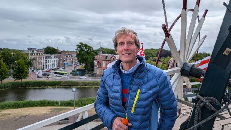 Een man in een blauwe winterjas staat op een platform naast een molen, met uitzicht op een stad met bomen en gebouwen; op de achtergrond is een rivier te zien.