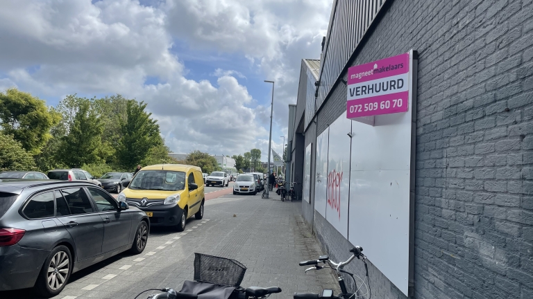 Straatbeeld met verschillende auto's geparkeerd langs de weg naast een grijs gebouw. Aan het gebouw hangt een bord van "magneet makelaars" met de tekst "VERHUURD" en een telefoonnummer. Er zijn fietsen geparkeerd langs de muur en de lucht is deels bewolkt.
