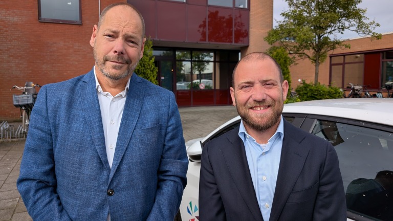 Twee mannen in pakken poseren voor een gebouw met rode bakstenen en grote ramen; op de achtergrond staat een fiets naast een auto.
