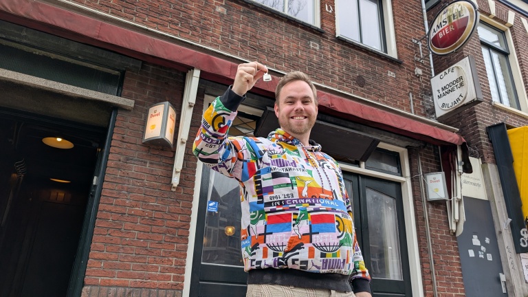Man in a colorful hoodie smiling and holding a key in front of a brick building with a bar sign.