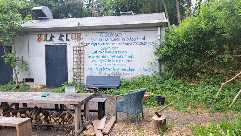 Schuur in een bosrijke omgeving met een kleurrijk bord "BUK KLUB" boven de deur en een blauwe tekst op de muur. Voor de schuur staan een houten tafel met houtstapels eronder, een stoel en een hakblok met een bijl. Omringd door bomen en struiken.