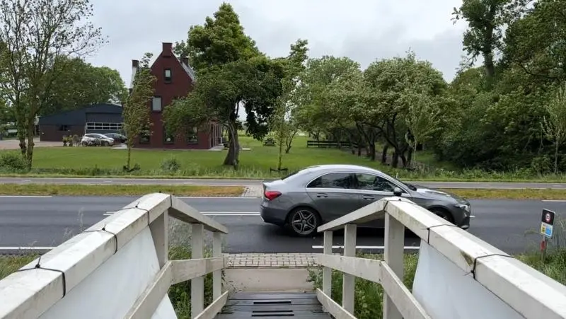 Trap leidt naar een weg met een passerende grijze auto; aan de overkant is een rode bakstenen gebouw met groen gras en bomen zichtbaar.