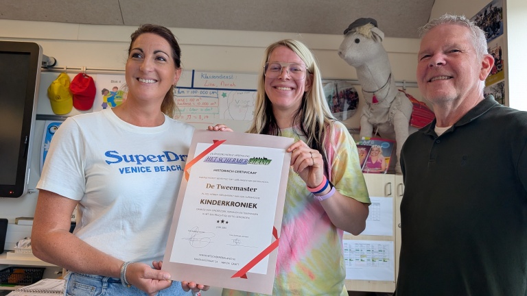 Drie mensen poseren met een certificaat in een klaslokaal, terwijl een knuffeldier op de achtergrond staat.