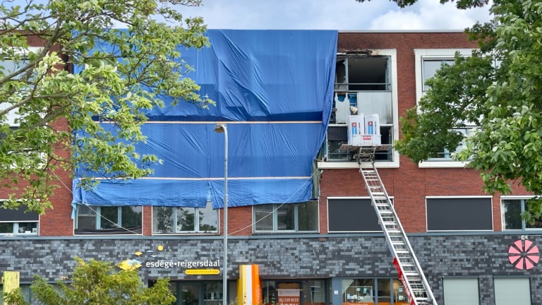 Een gebouw met deels bedekte gevel door een groot blauw doek; er zijn bouwmaterialen zichtbaar op een platform, ondersteund door een schuine ladder. Bomen flankeren het beeld.