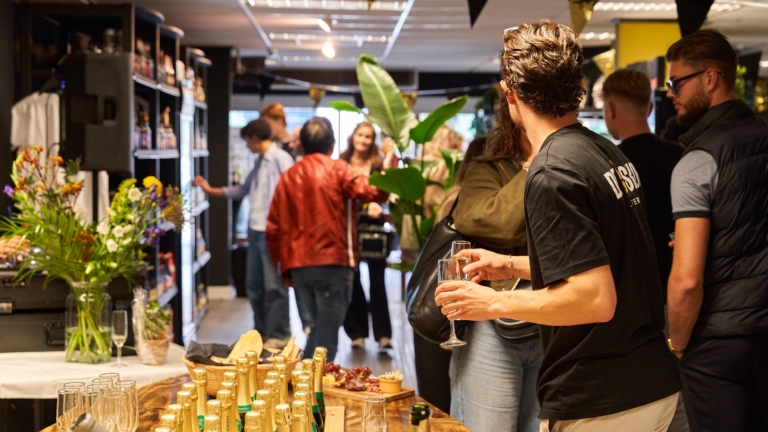 Een groep mensen verzameld in een winkelachtige ruimte met flessen champagne en glazen op een tafel, terwijl ze ontspannen en praten tijdens een evenement; er hangen feestelijke vlaggen en er zijn bloemen als decoratie.