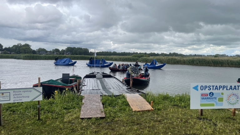 Een aanlegsteiger aan een rivier met meerdere boten waarop mensen zitten onder een bewolkte hemel, omgeven door grasachtige oevers. Een bord met de tekst 'OPSTAPPPLAATS' en een routebeschrijving is zichtbaar op de voorgrond.