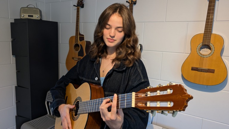 Een meisje speelt gitaar in een kamer met meerdere gitaren aan de muur.