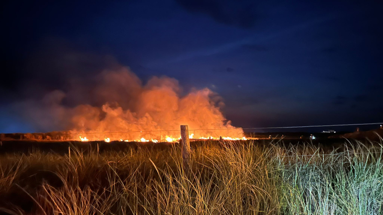 Een nachtelijk landschap met een veldbrand die oranje rook en vlammen produceert, gefotografeerd vanuit het gras op de voorgrond.