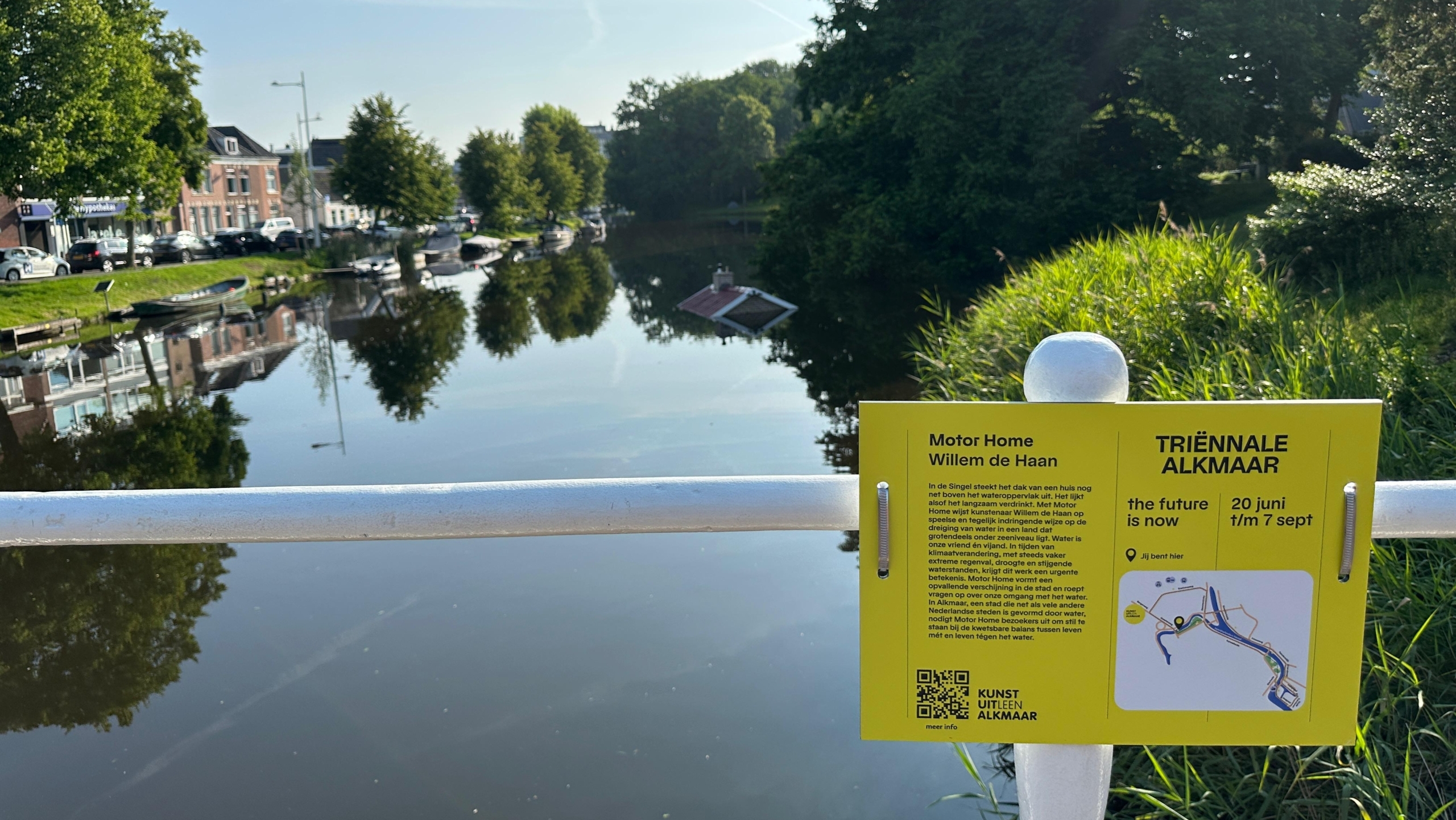 Uitzicht op een kanaal met bomen en gebouwen aan de zijkanten, en enkele boten aangemeerd. Op de voorgrond is een geel bord bevestigd aan een witte reling met informatie over 'Motor Home' van Willem de Haan voor de Triënnale Alkmaar, met data en locatie-informatie.
