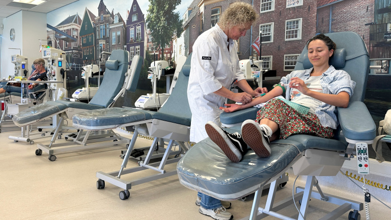 Een vrouw doneert bloed in een donatiecentrum, zittend in een comfortabele stoel terwijl een verpleegkundige een naald inbrengt. Op de achtergrond is nog een persoon te zien die bloed doneert, met een muurschildering van Nederlandse grachtenhuizen als decor.