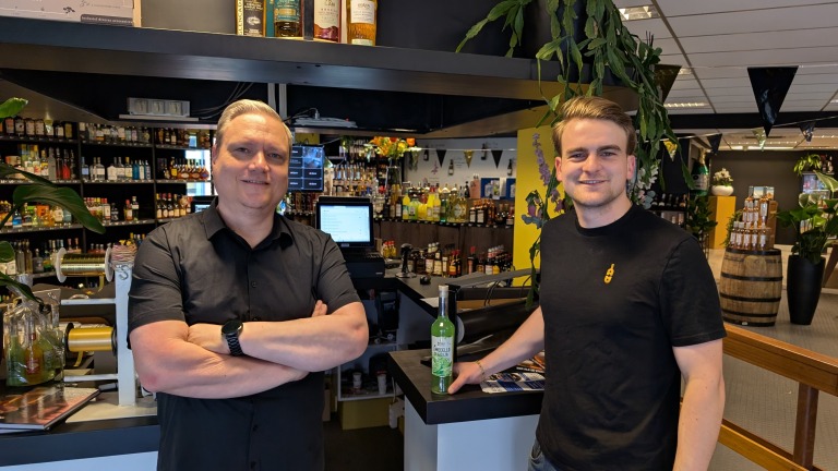 Twee mannen staan in een drankenwinkel voor een balie, omringd door flessen sterke drank en decoratieve planten.