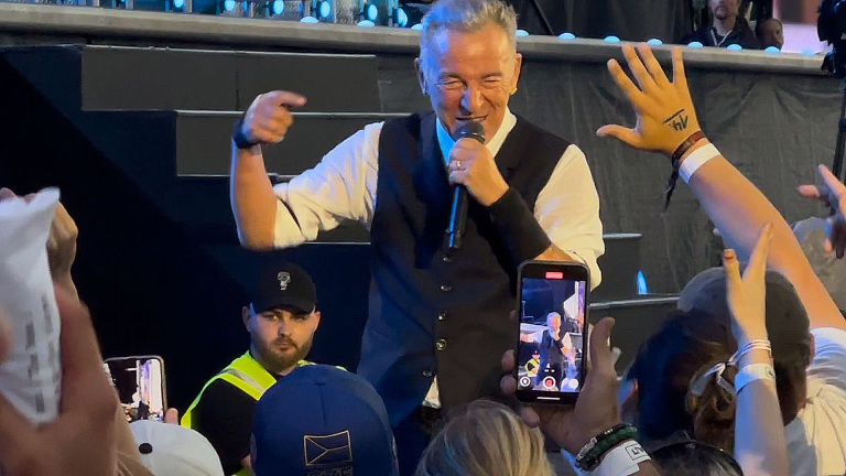 Een zanger met een microfoon in zijn hand staat dicht bij het publiek op een podium, terwijl toeschouwers hun handen naar hem uitstrekken en foto's maken met hun telefoons.