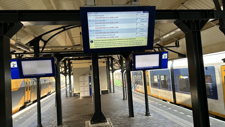 Een leeg treinstation met een NS-trein op de achtergrond en meerdere informatieschermen met berichten over treinannuleringen vanwege een staking.