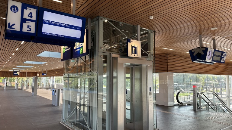 Modern treinstation met een glazen lift, richtingbordjes voor perrons 4 en 5, en een roltrap. Er zijn digitale schermen met treininformatie en een klok in een interieur met houten plafond.