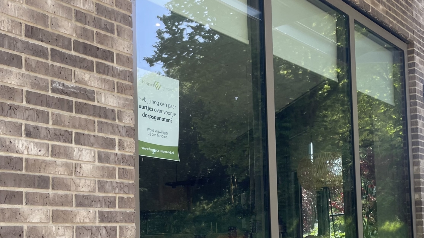 Zijaanzicht van een gebouw met bakstenen muren en grote ramen, met een poster op het raam. Op de voorgrond is een metalen paneel met het cijfer 2 zichtbaar, en op de achtergrond is een auto geparkeerd naast het gebouw, omringd door groen.