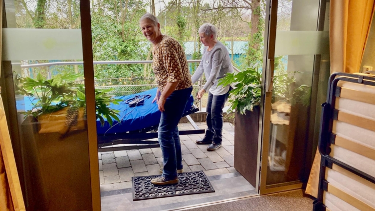 Twee oudere vrouwen staan glimlachend op een terras met planten aan weerszijden, terwijl een opklapbaar bed op de voorgrond zichtbaar is.