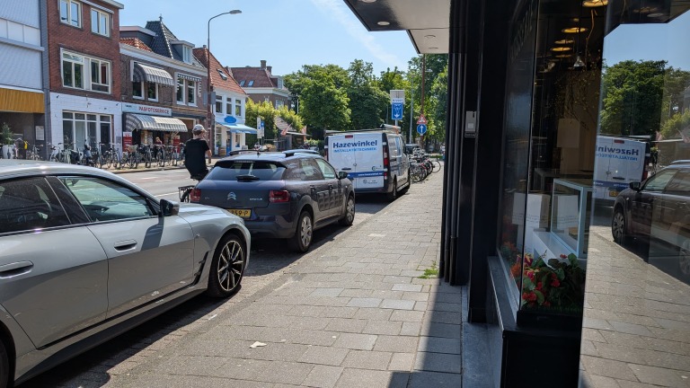 Straatbeeld met geparkeerde auto's langs de stoep en een rij met fietsen, naast enkele winkels, waaronder een pasfotoservice.