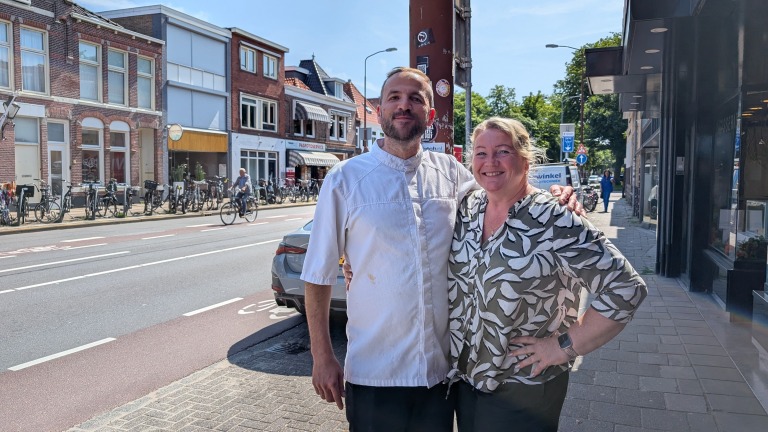 Twee mensen staan dichtbij elkaar en glimlachen terwijl ze op een zonnige straat in een stedelijke omgeving poseren, omringd door fietsen en passerende mensen.