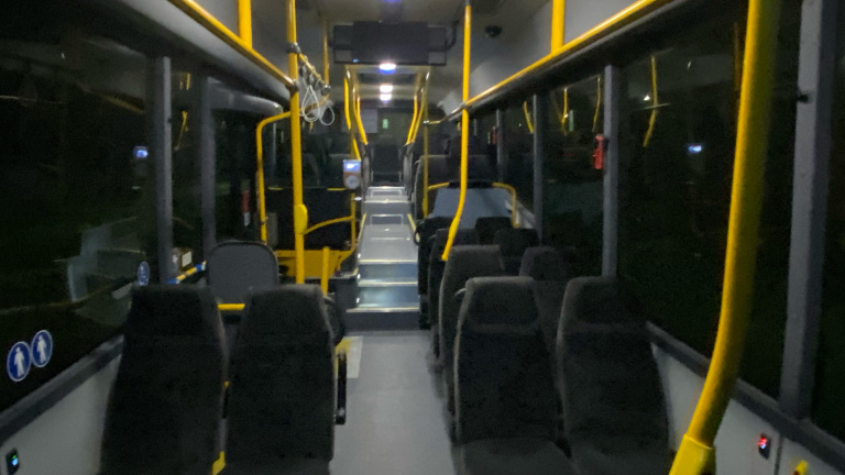 Het interieur van een lege stadsbus met zwart-grijze stoelen, gele relingen en een donkere buitenomgeving zichtbaar door de ramen.