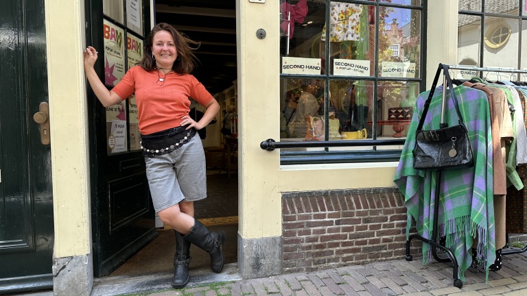 Een vrouw in een rode trui en grijze shorts staat in de deuropening van een tweedehandswinkel. Naast haar hangt kleding aan een rek.