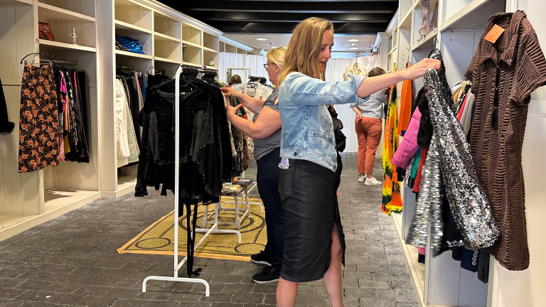 Twee vrouwen winkelen in een kledingwinkel, waarbij de voorste vrouw een sprankelende zilveren jurk bekijkt, omgeven door rekken met diverse gekleurde kledingstukken in een lichte en ruime boetiek.