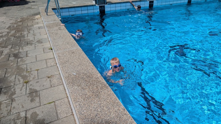 Twee kinderen met zwembrillen spelen in een buitenzwembad, terwijl een derde persoon op de achtergrond zwemslagen uitvoert.