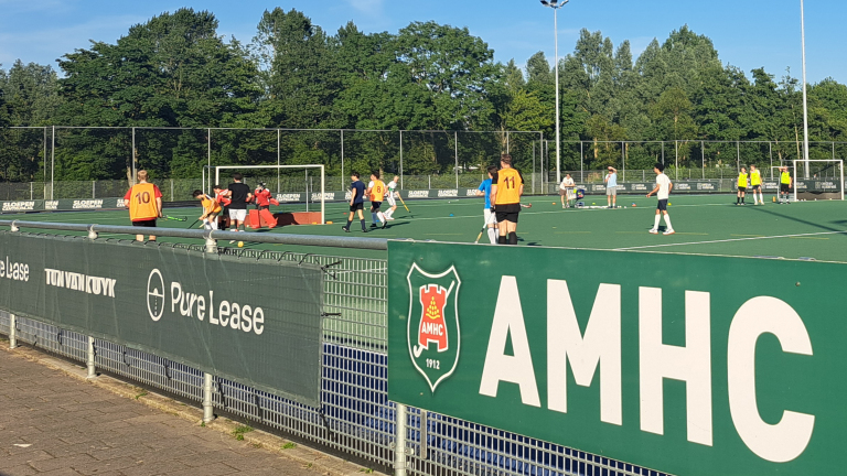 Hockeyspelers trainen op een veld van de AMHC club, met spelers in oranje hesjes en anderen in witte en blauwe shirts. Op de voorgrond is een hek met reclameborden zichtbaar, waaronder het logo van AMHC en het jaartal 1912. Op de achtergrond zijn bomen en een heldere lucht te zien.
