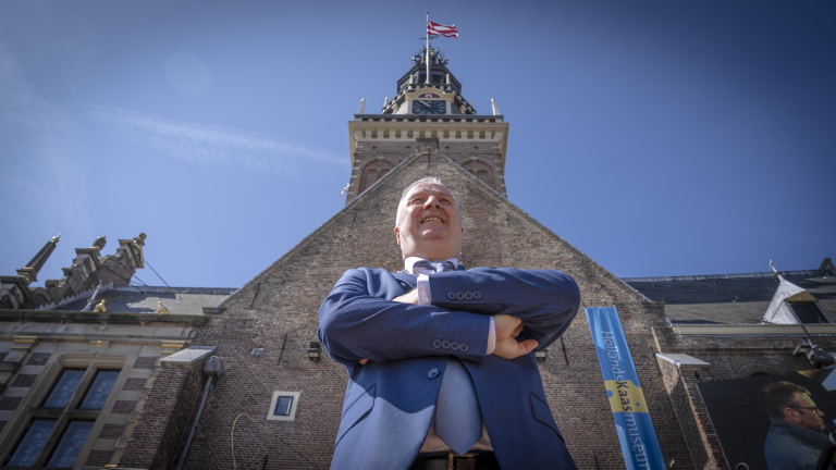 Man in blue suit with arms crossed, standing confidently in front of a historic brick building with a clock tower and flag on top, under a clear blue sky.
