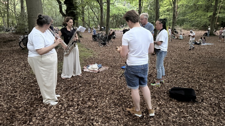 Een groep mensen speelt muziek in een bos, met een vrouw die een saxofoon bespeelt en een andere die een dwarsfluit bespeelt, omringd door anderen die aandachtig kijken. De bosgrond is bedekt met bladeren en er staan enkele fietsen in de achtergrond.